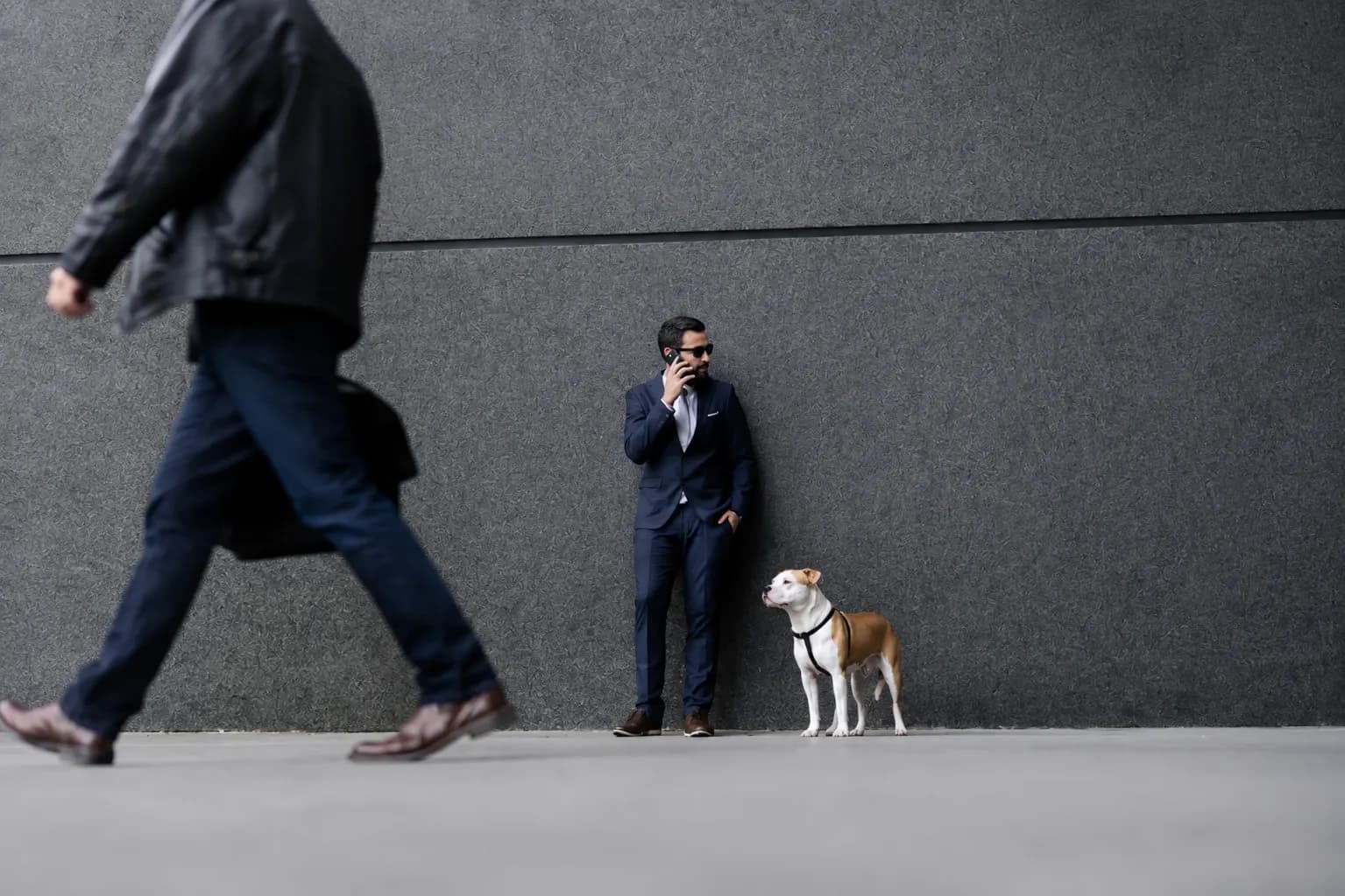 Profissional de terno ao telefone encostado em parede cinza, com seu cão ao lado, em calçada de centro urbano